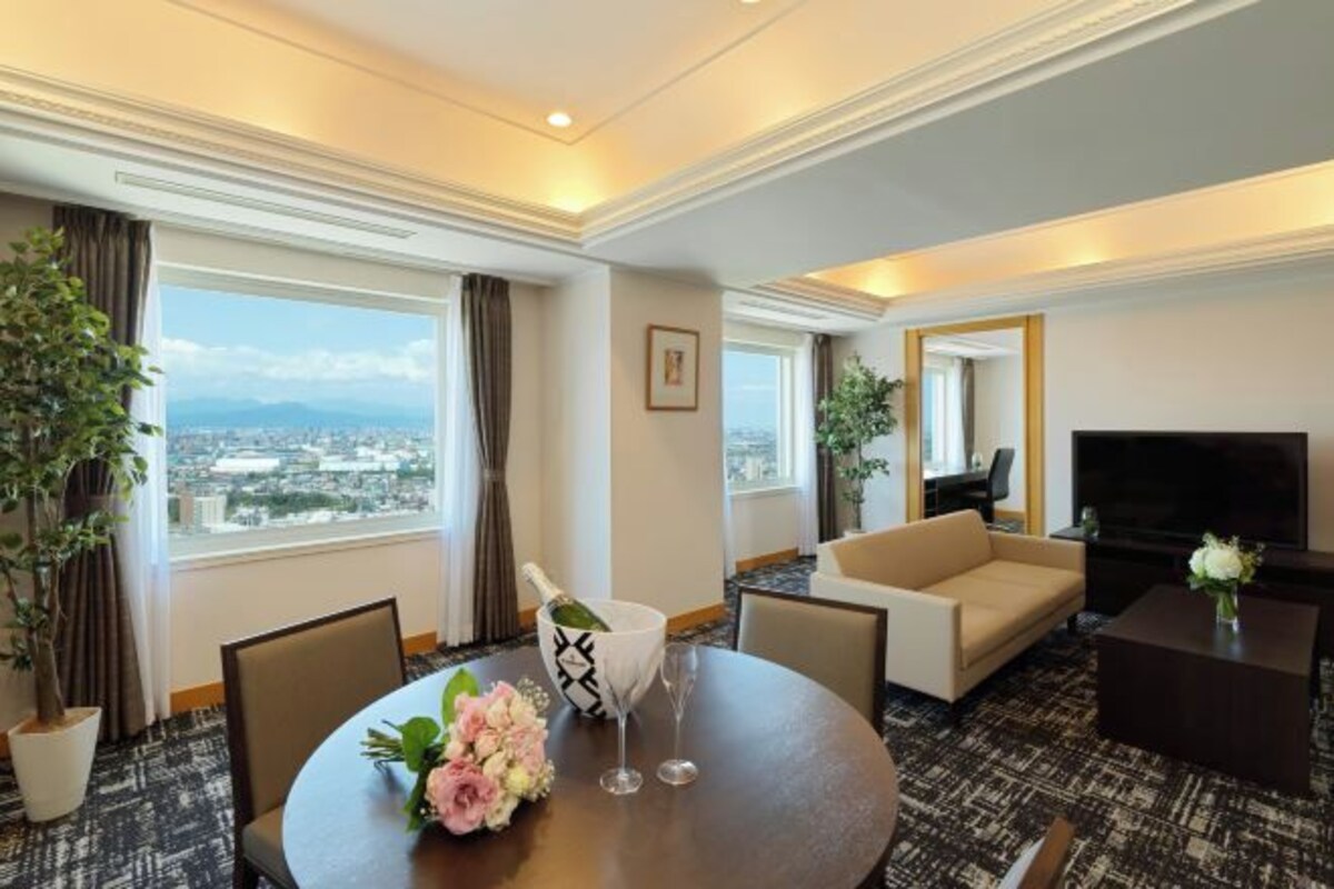 A hotel room with a round dining table, a sofa, a TV, and a large window offering a city view.