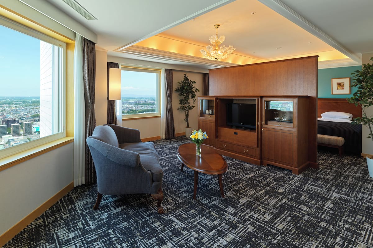 A hotel room with a large window offering a city view, a sofa, a coffee table, a TV, and a bed.