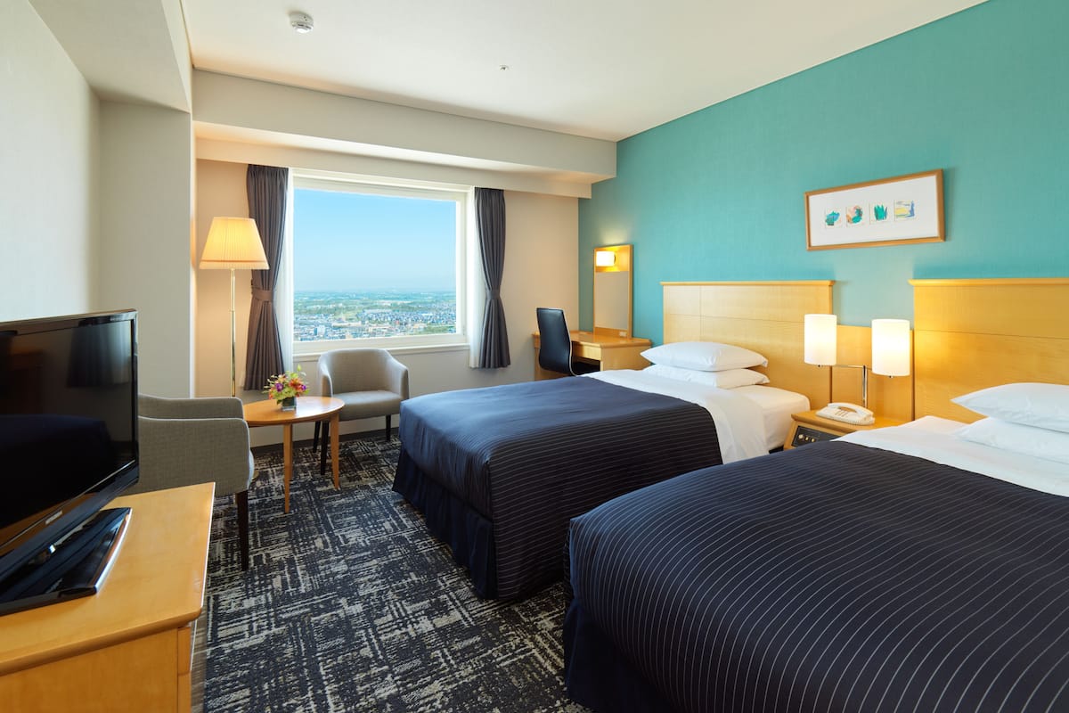 A hotel room with two beds, a desk, a TV, and a view of the city.