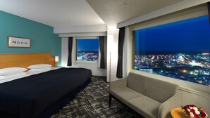 Ein Hotelzimmer mit einem Bett, einer Couch, einem kleinen Tisch mit Blumen und einem großen Fenster mit Blick auf die nächtliche Stadt.