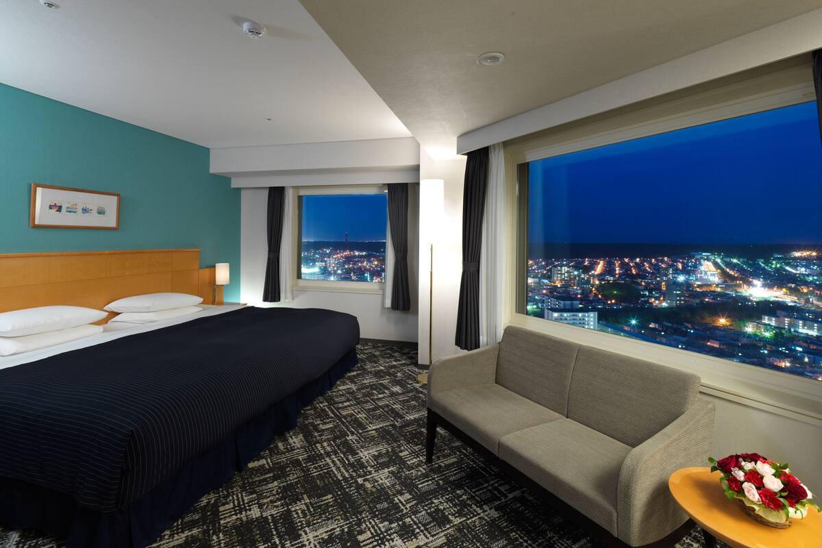 A hotel room with a bed, a sofa, a small table with flowers, and a large window offering a city view at night.