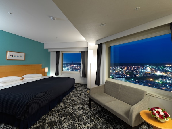Habitación de hotel con cama, un sofá, una mesita con flores y un ventanal grande que ofrece vista a la ciudad de noche.