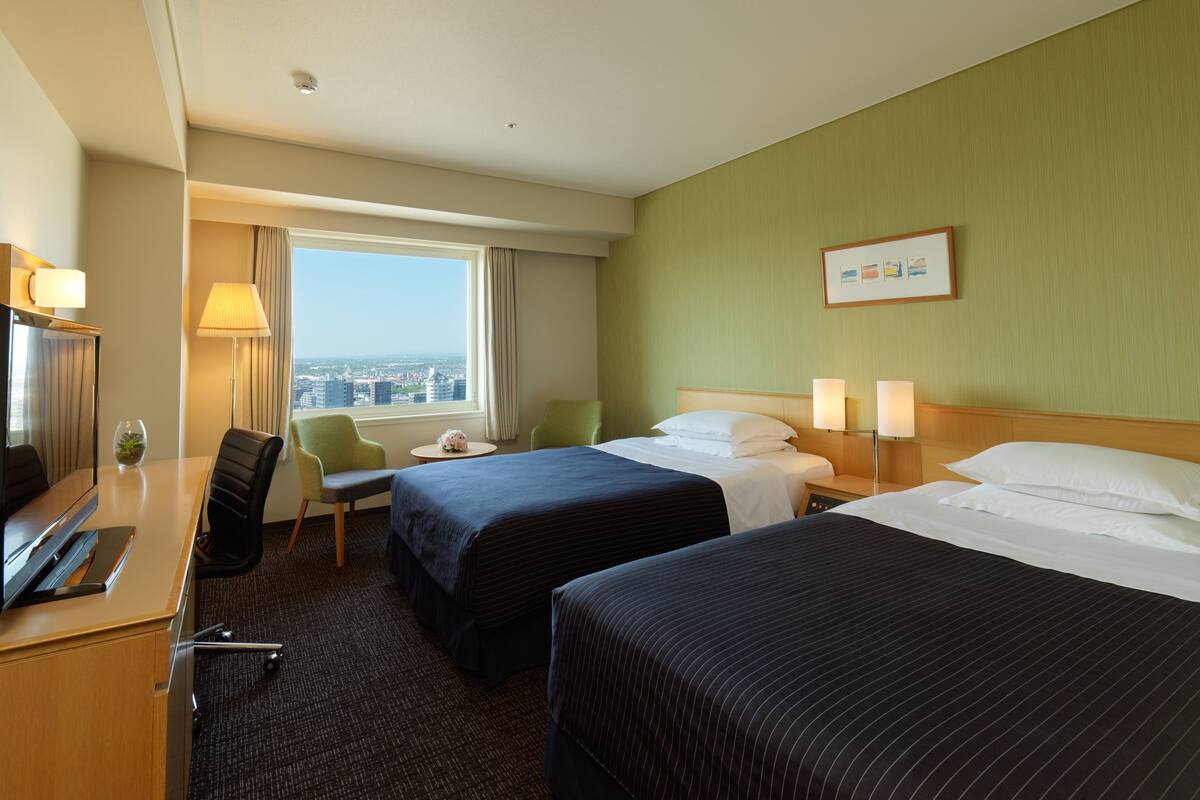 A hotel room with two beds, a desk, a chair, a TV, and a window with a city view.