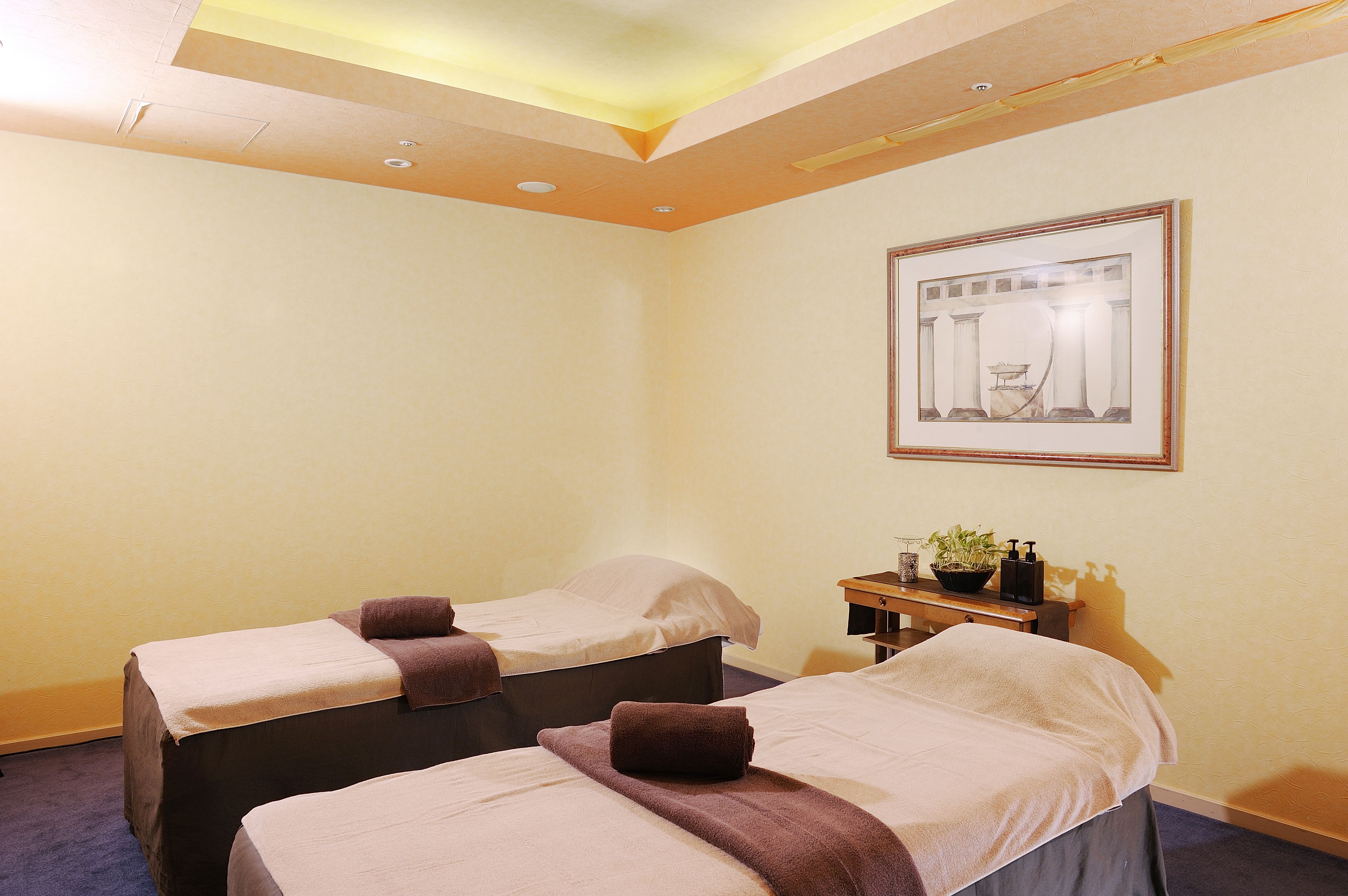 A hotel room with two single beds, a small table with a plant, and a framed picture on the wall.