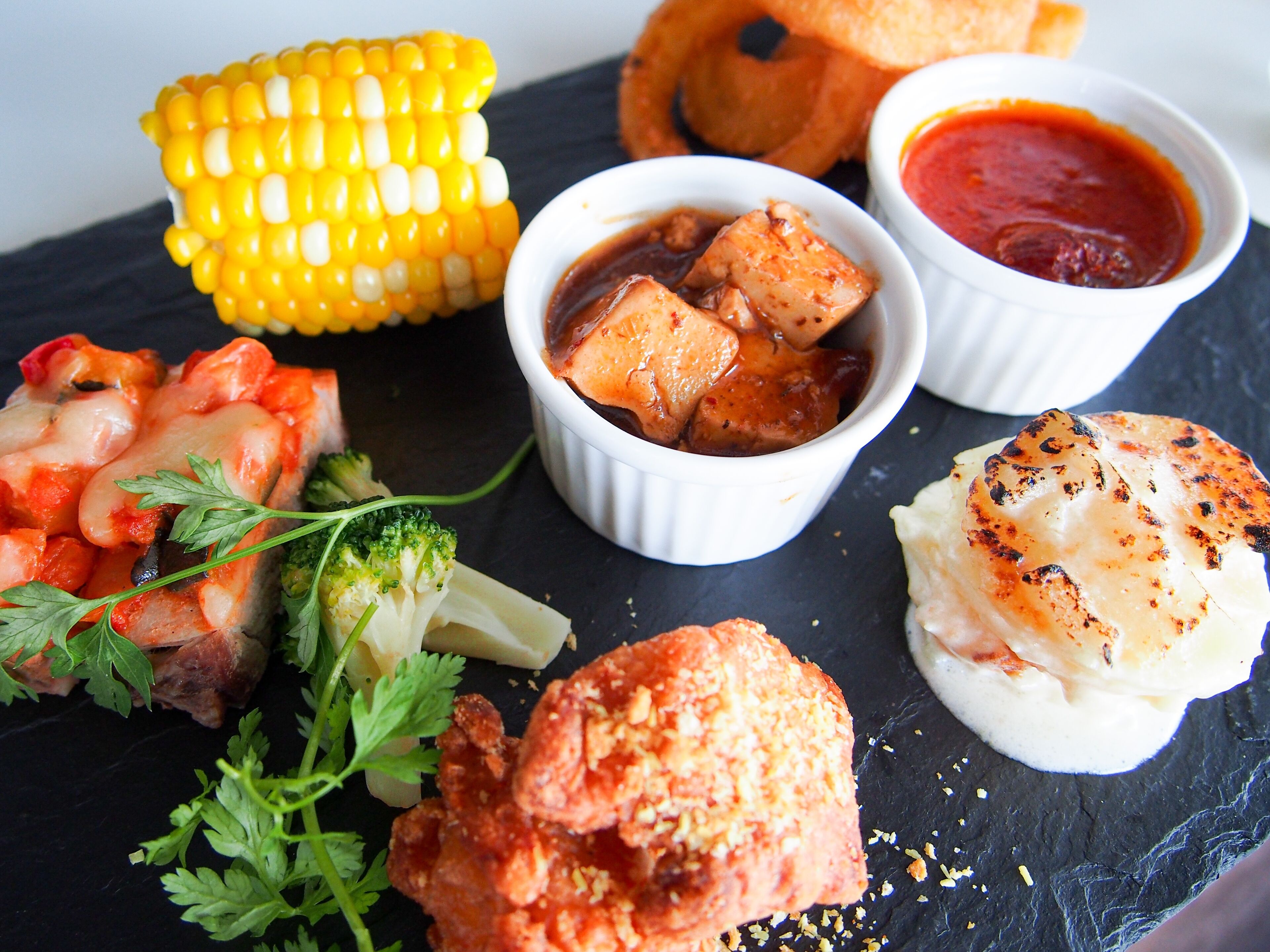 A variety of dishes including grilled chicken, corn on the cob, and a piece of fish, served on a slate board.