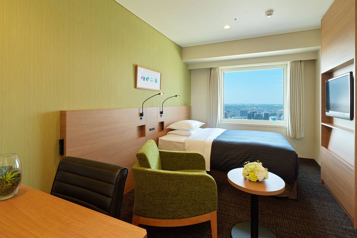 A hotel room with a bed, a desk, a chair, a small table with flowers, a TV, and a window with a city view.