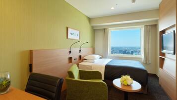 Una habitación de hotel con cama, escritorio, silla, una pequeña mesa con flores, televisión y ventana con vista a la ciudad.