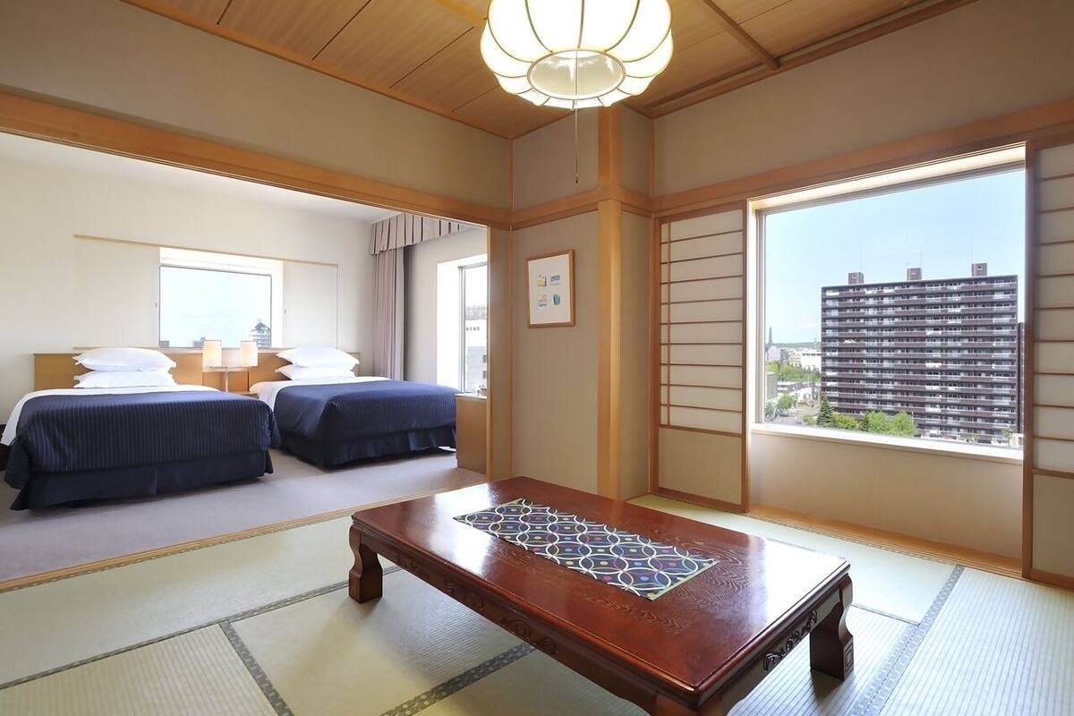 A traditional Japanese-style room with tatami flooring, a low wooden table, two beds with blue bedding, and a large window offering a city view.