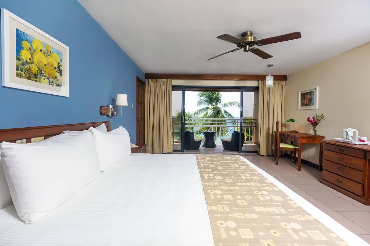 A hotel room with a bed, a desk, a chair, a ceiling fan, and a balcony with a view of palm trees.