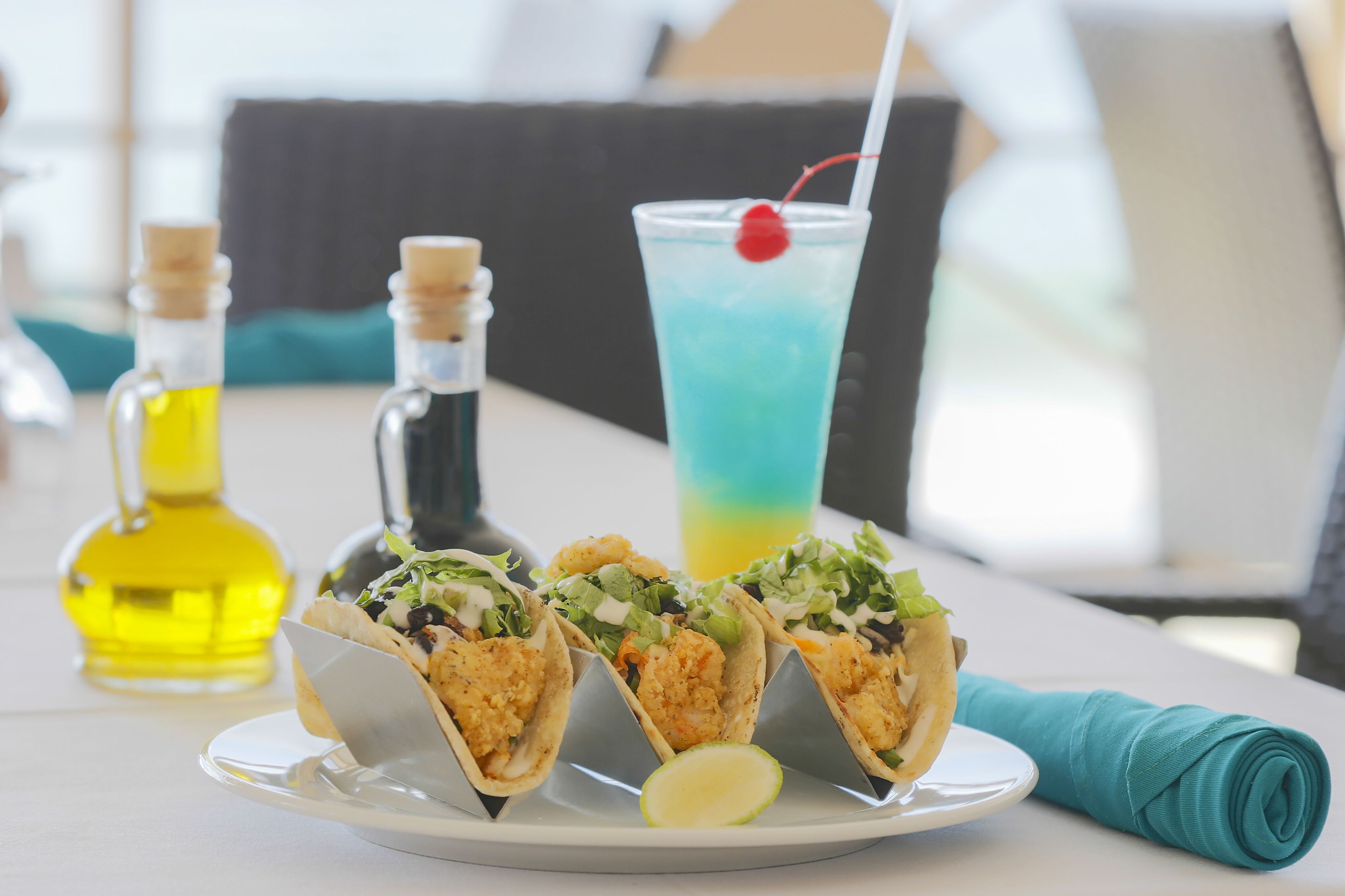 Three seafood tacos on a plate with a lime wedge, a cocktail with a cherry, and two bottles of oil on a table.