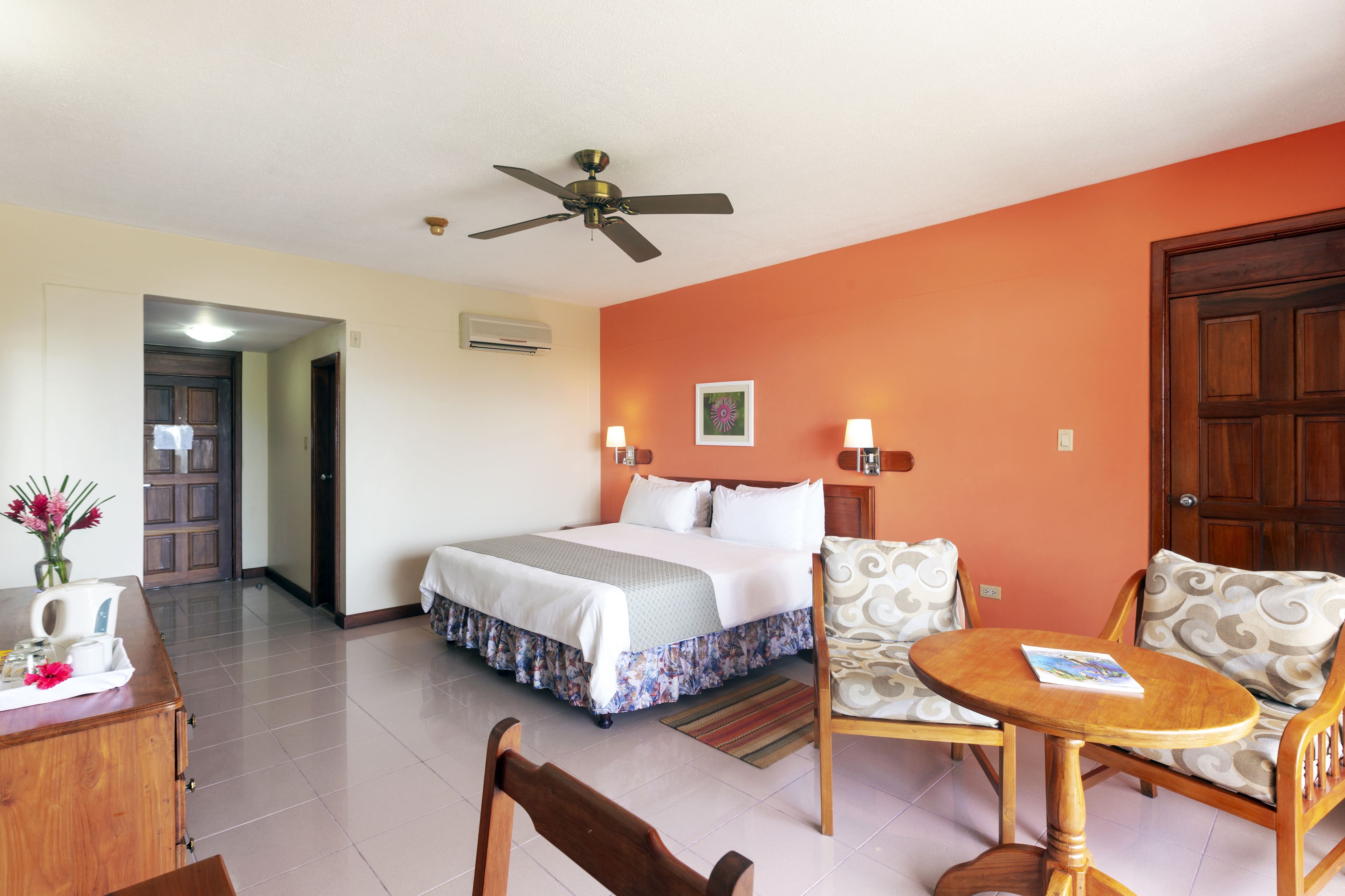 A hotel room with a bed, a wooden table, two chairs, a ceiling fan, and a door.