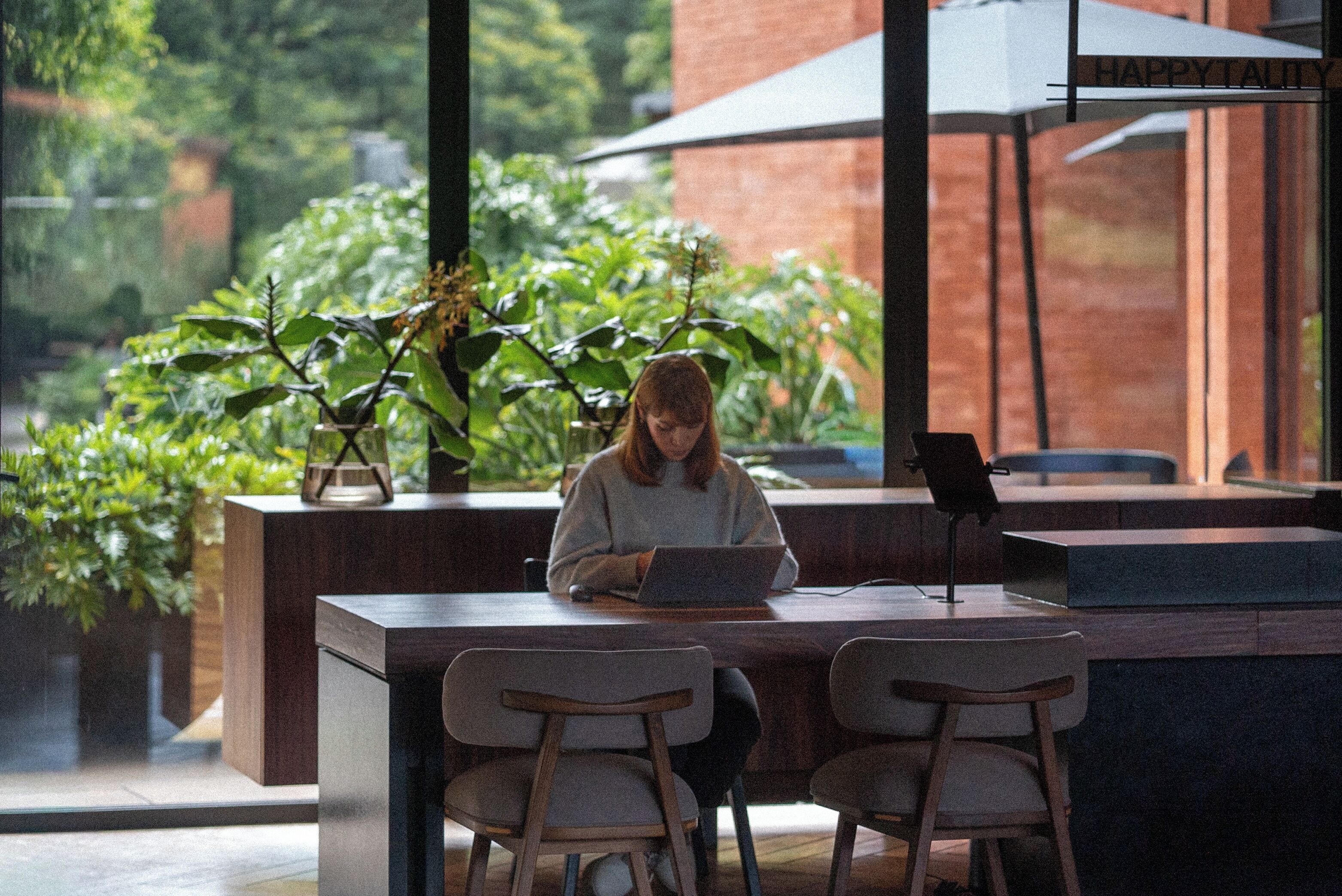 A person working at a wooden table with a laptop, surrounded by potted plants and a brick wall.