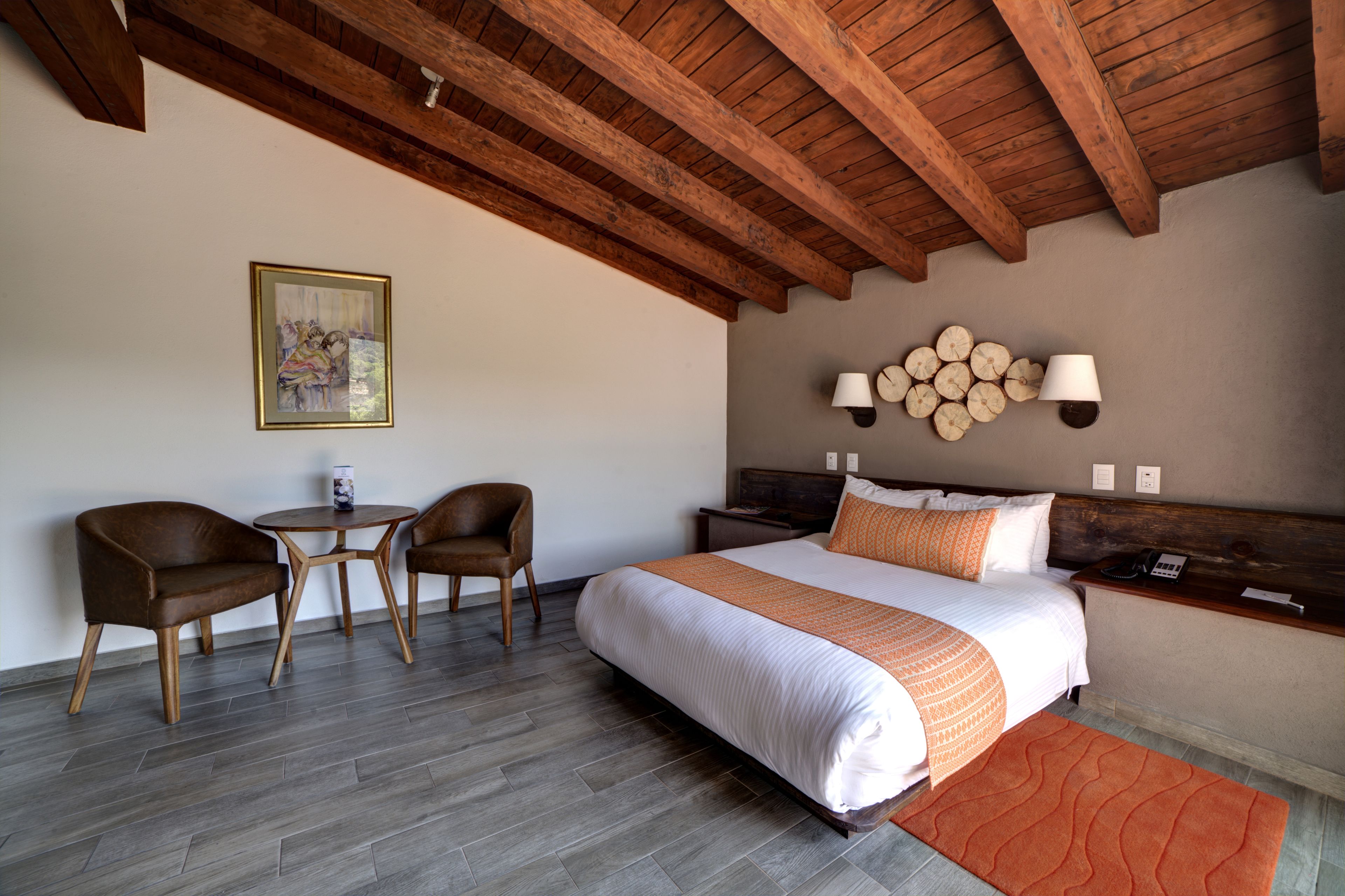 A hotel room with a wooden ceiling, a bed with white and orange bedding, two chairs, a small table, and a wall-mounted lamp.