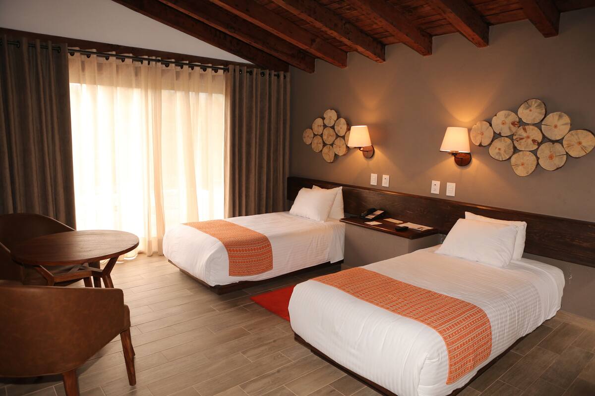 A hotel room with two beds, a wooden table, and a chair. There are wall-mounted lamps and a decorative wall piece.