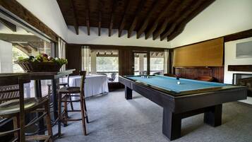 A pool table with a green felt surface, wooden chairs, a wicker basket with fruits, and a white tablecloth.