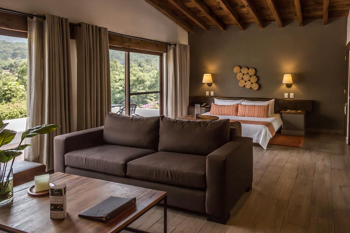 A modern hotel room with a large window, a sofa, a bedside table, a bed with pillows, and a wall clock.