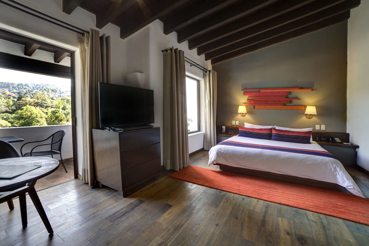 A modern hotel room with a large bed, a flat-screen TV, a dining table, and a view of the outdoors.
