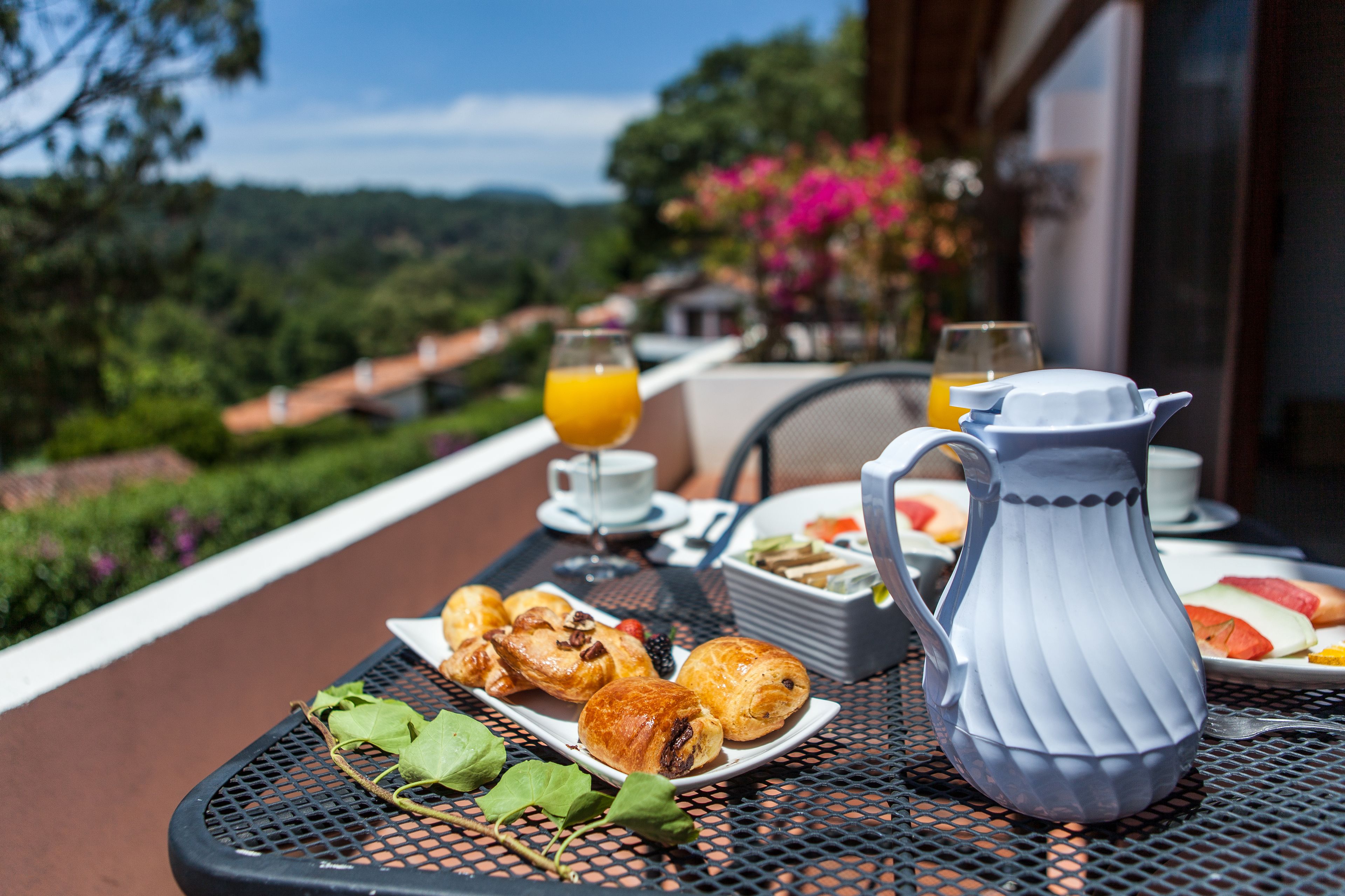 A table with breakfast items including croissants, a pitcher, and a glass of orange juice, set against a scenic view.
