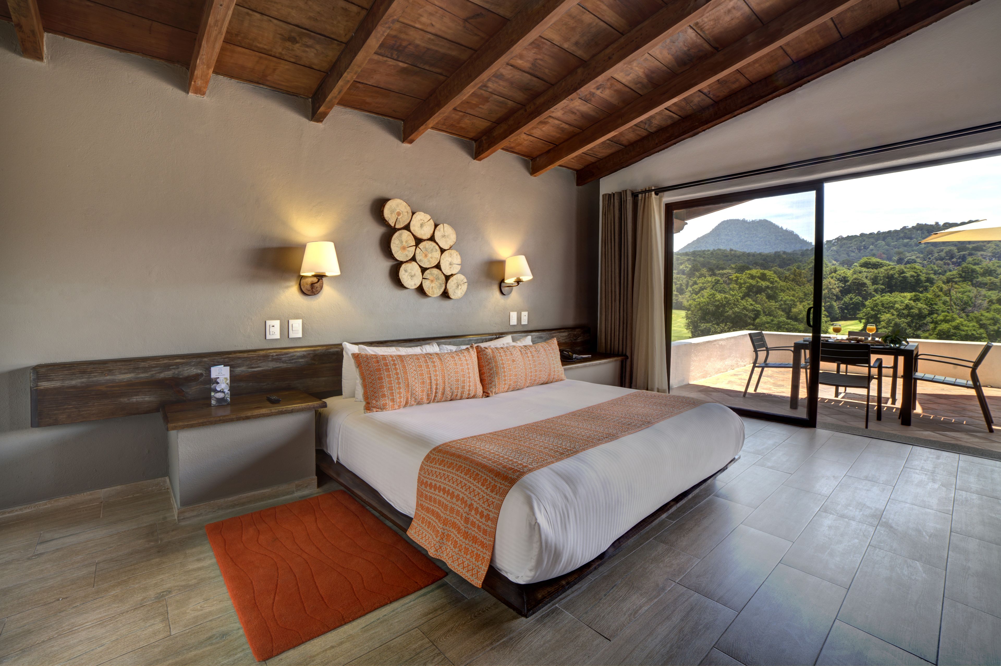 A bedroom with a large bed, wooden headboard, bedside tables, and a view of a mountainous landscape through a sliding glass door.