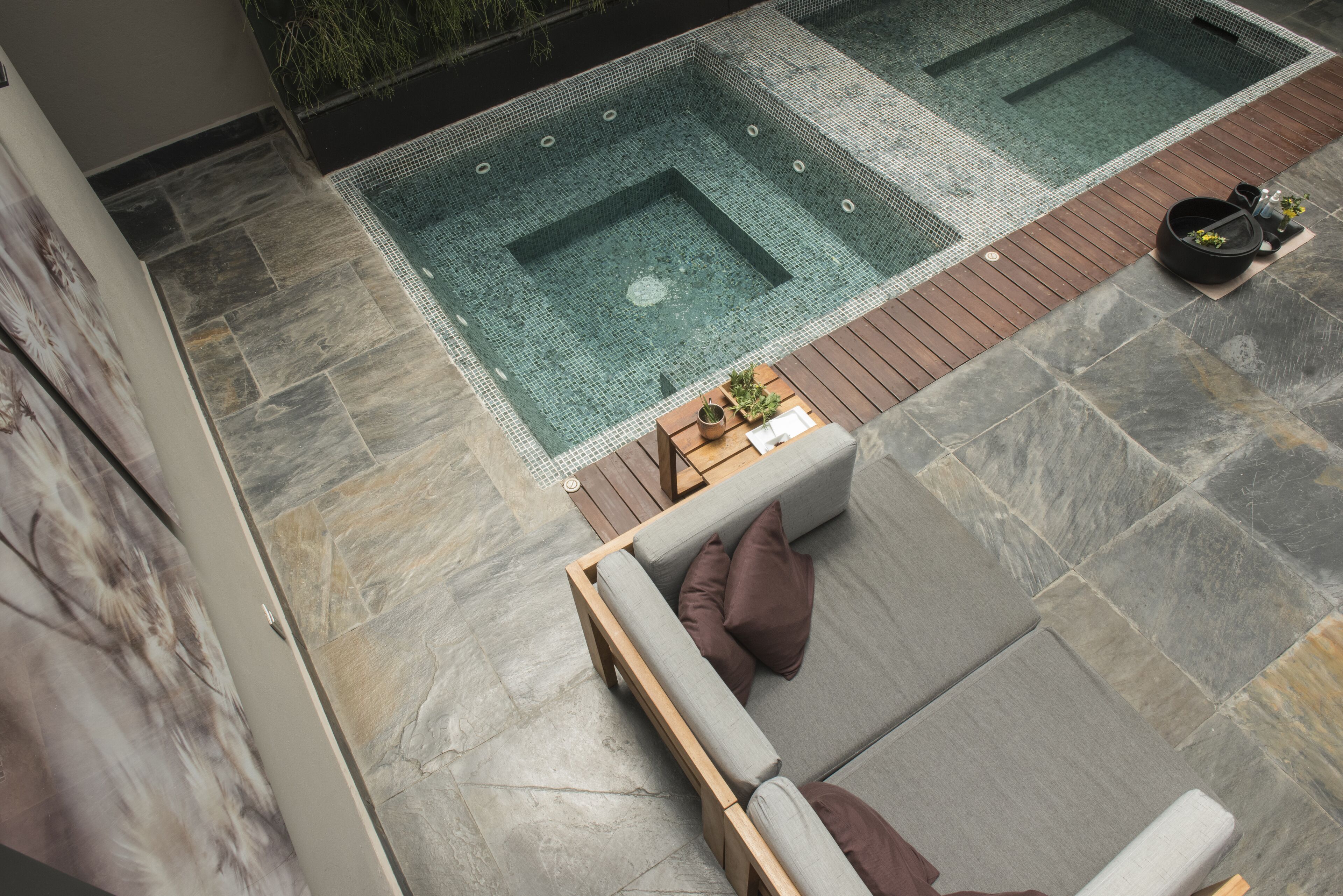 A modern outdoor pool area with a wooden deck, a seating area with cushions, and a stone-tiled floor.