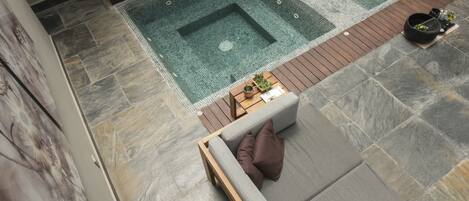 A modern outdoor pool area with a wooden deck, a seating area with cushions, and a stone-tiled floor.