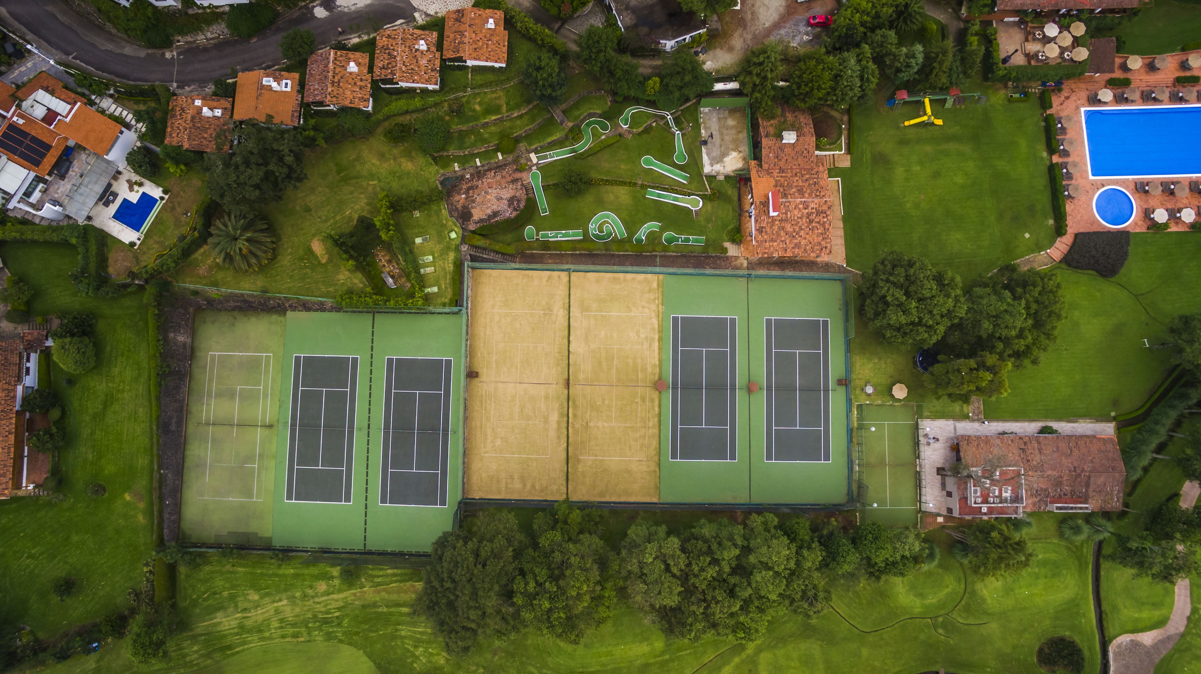 An aerial view of a residential area with a large tennis court, a swimming pool, and surrounding greenery.