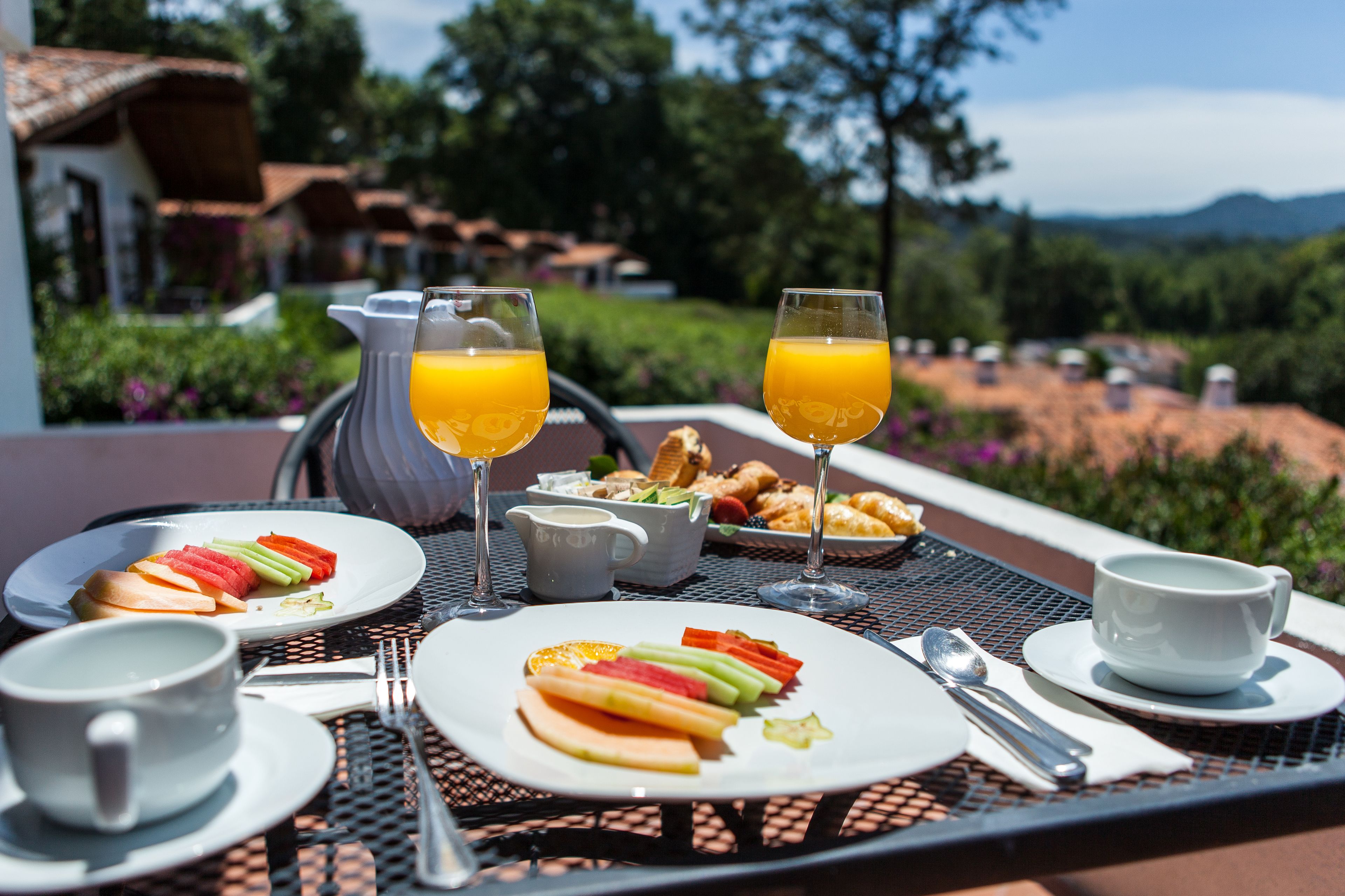 A breakfast table with plates of fruit, glasses of orange juice, a pitcher, and cups.