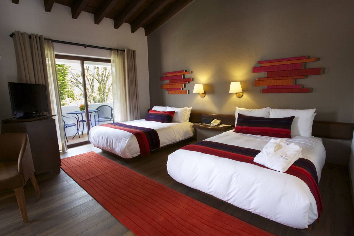 A hotel room with two beds, a desk with a TV, and a balcony with a chair.