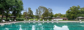 Una piscina con agua cristalina, rodeada de sillones de descanso y sombrillas, con árboles y un edificio de fondo.