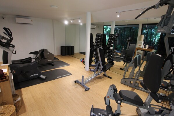 A gym with exercise equipment, a mirror, and a view of trees through the windows. - Paradise Beach Resort Samui (Koh Samui)