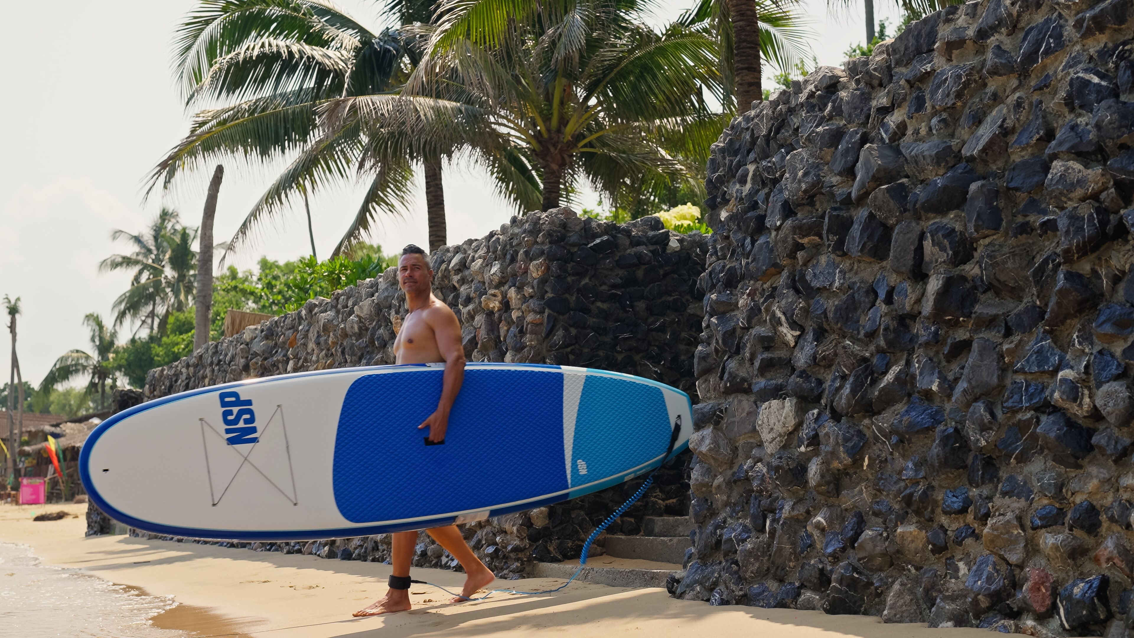 A man carrying a blue and white surfboard with "NSS" on it, walking on a sandy beach with palm trees and a stone wall in the background.