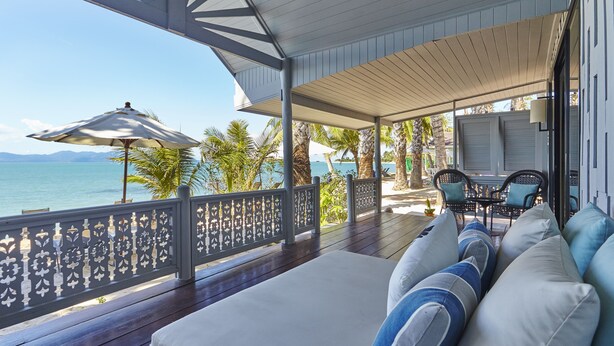 Un espace extérieur abrité avec des places assises, offrant une vue sur la mer, une terrasse en bois et un canapé blanc avec des coussins bleus.