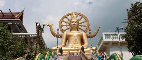 A large golden statue of a seated figure with a wheel at its feet, surrounded by ornate architecture and a staircase leading up to it.