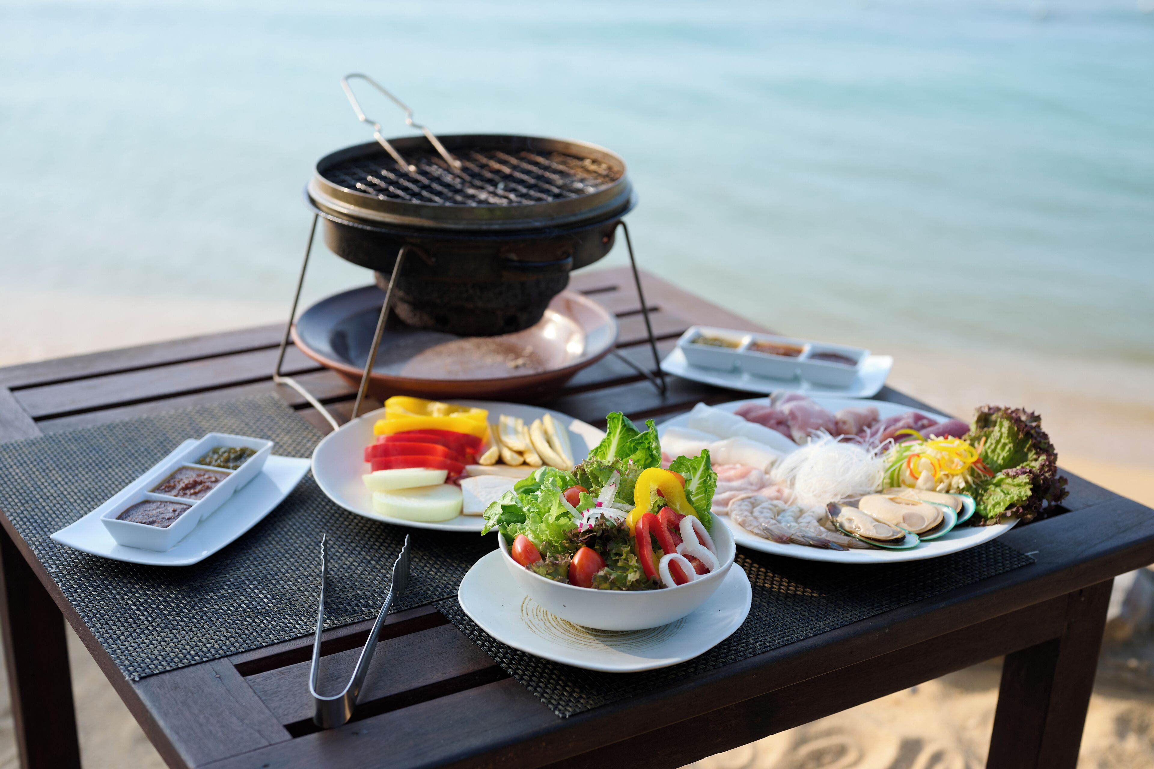 Une table dressée sur la plage, où sont disposés divers aliments, notamment une salade, des légumes coupés et des fruits de mer, pour un repas.