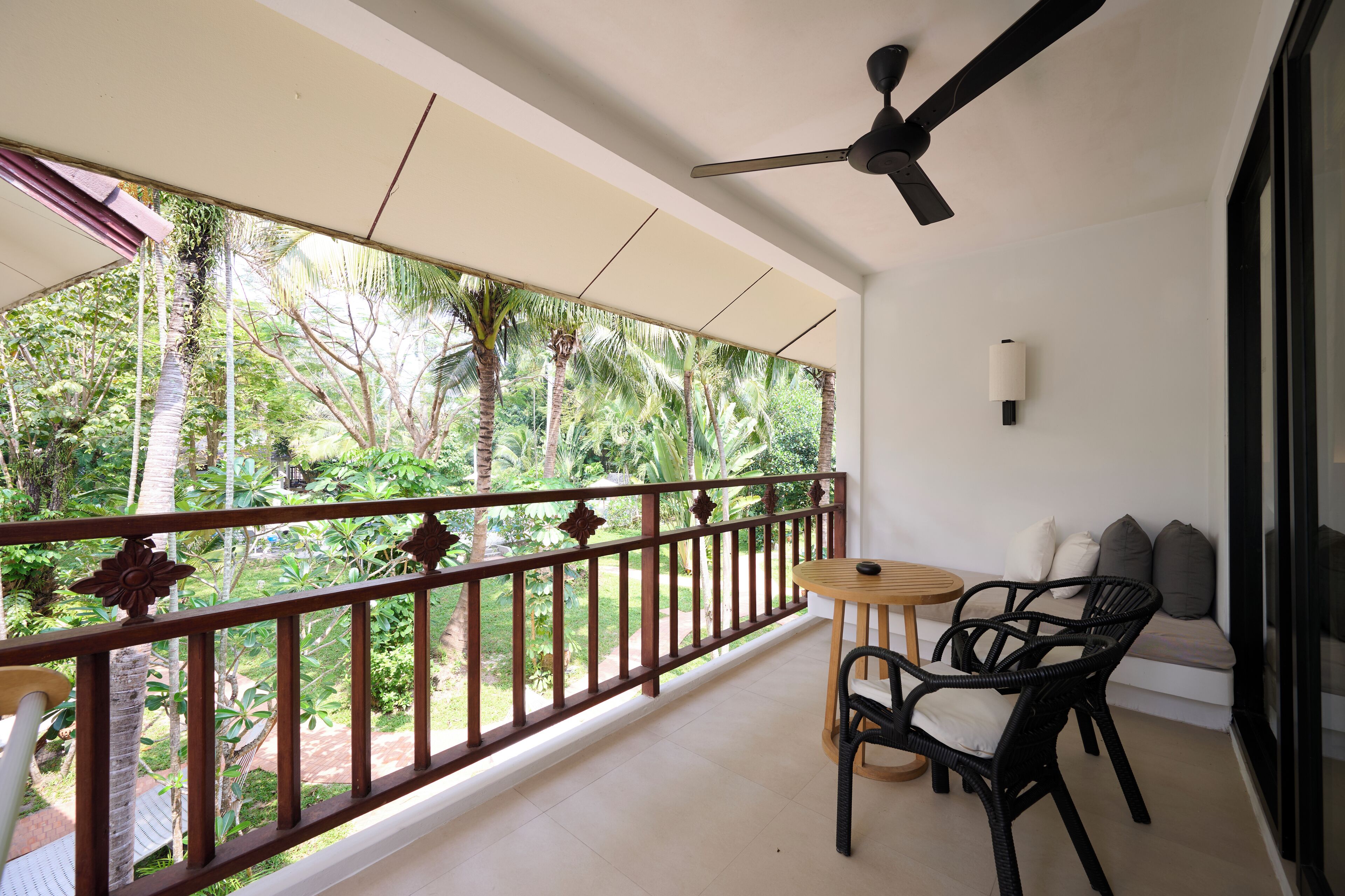 Un balcon donnant sur une végétation tropicale, un ventilateur de plafond et un coin salon avec une table et des chaises.