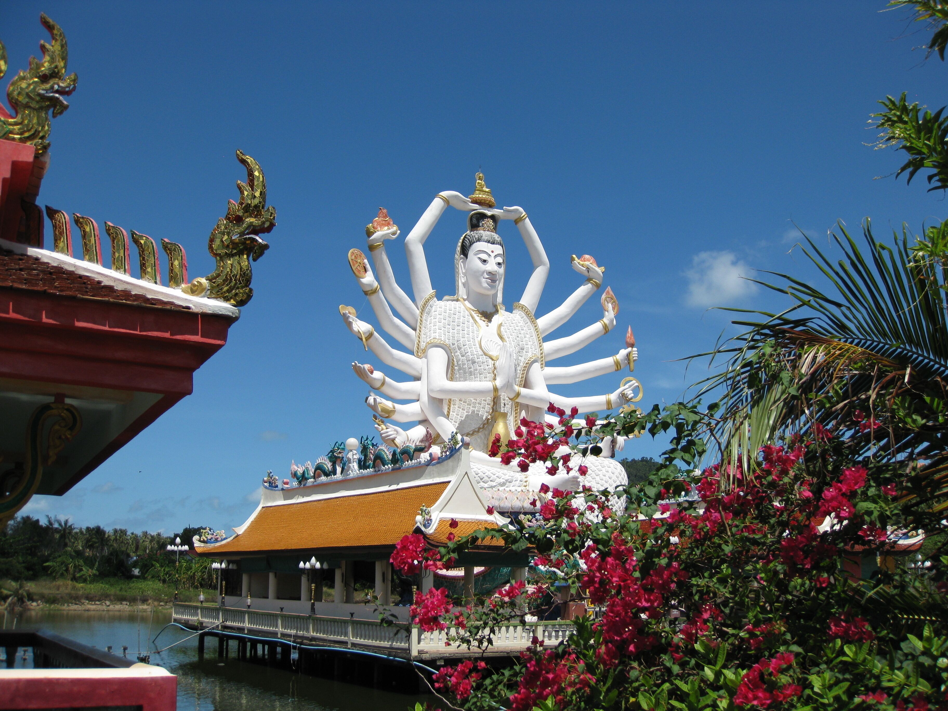 Eine große weiße, mehrarmige Statue mit goldener Kopfbedeckung, umgeben von üppigen rosa Blumen und einem traditionellen Gebäude mit gelbem Dach.
