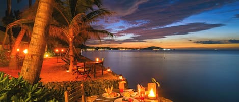Un área de comedor al aire libre con una mesa lista para dos, con vista a un cuerpo de agua tranquilo y un cielo al atardecer.