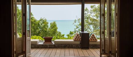 A room with a wooden floor, a cushioned bench, and a view of the sea and greenery.