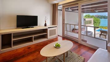 A modern living room with a flat-screen TV, wooden flooring, a small table with apples, and a balcony with a pool and scenic views.