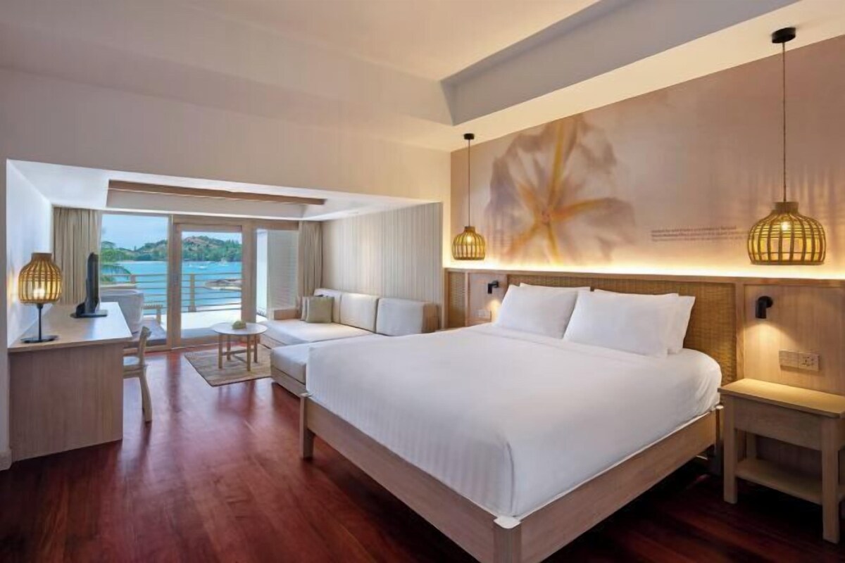 A hotel room with a large bed, a desk, a chair, and a view of the sea.