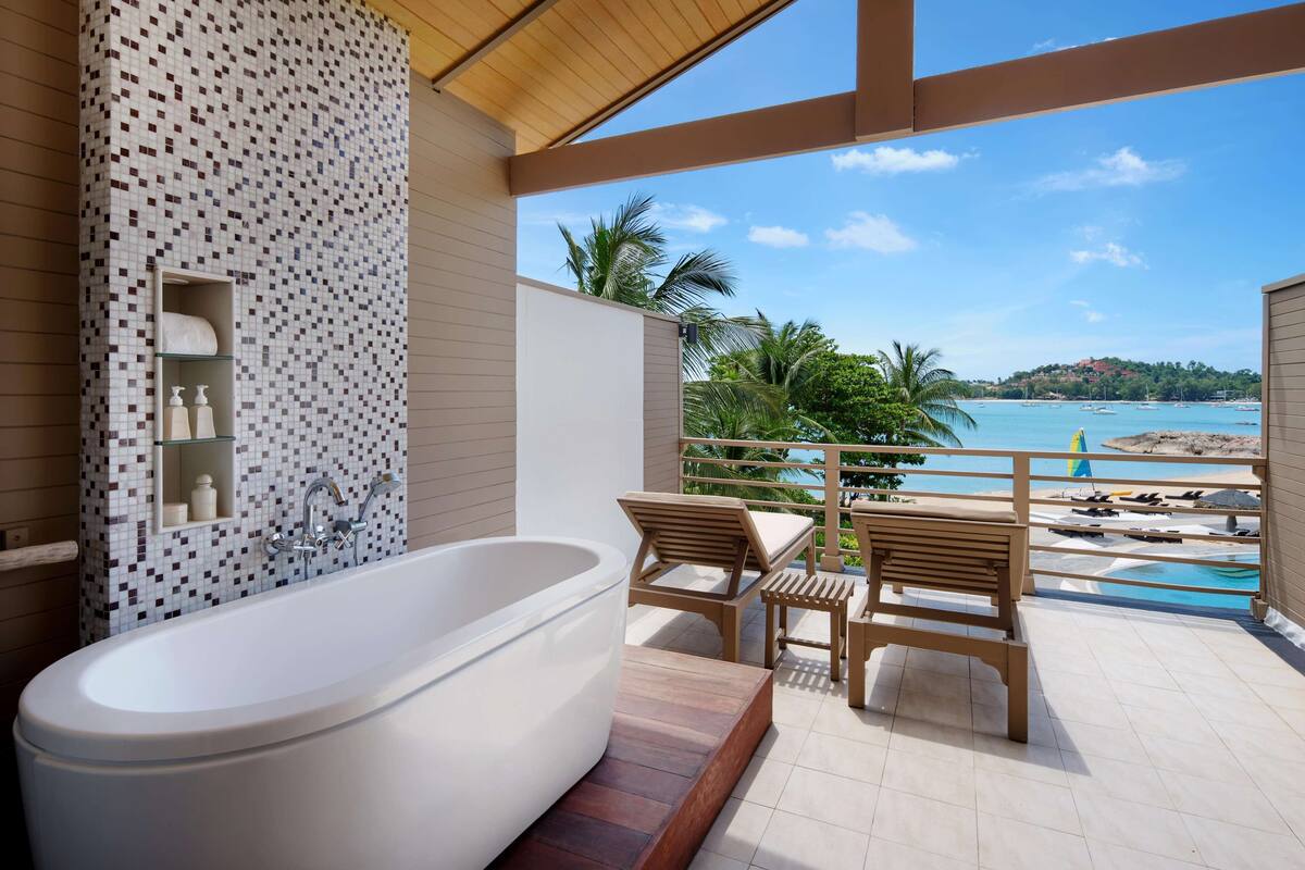 A modern bathroom with a freestanding bathtub, wooden chairs, and a view of the sea.