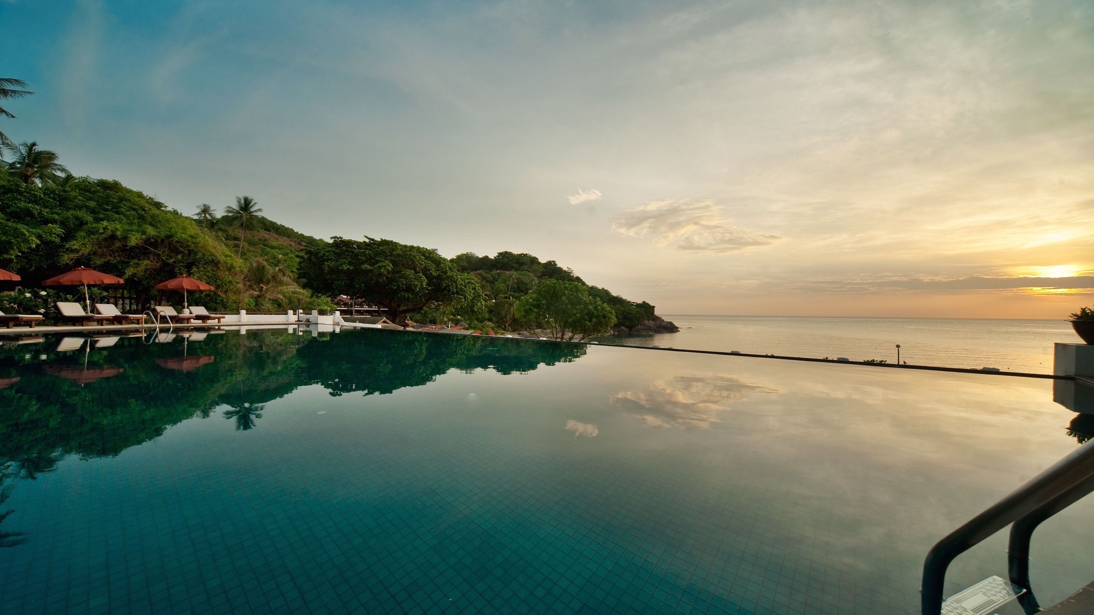 Piscina cristalina reflejando el atardecer, rodeada de sillones de descanso y sombrillas, con vista al mar y árboles.
