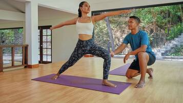 Una clase de yoga en una habitación con amplios ventanales, pisos de madera y colchonetas de yoga color púrpura.