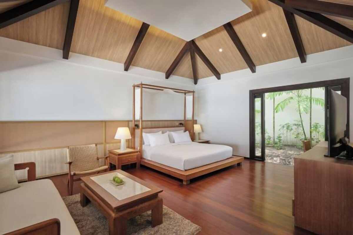 Un dormitorio con cama con dosel, suelo de madera, un escritorio y vistas a la vegetación a través de un ventanal.
