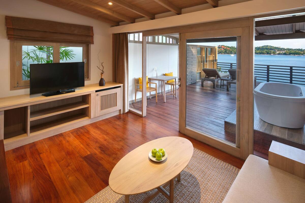 A modern living room with a wooden floor, a TV on a wooden console, a table with apples, and a balcony with a view of the sea.