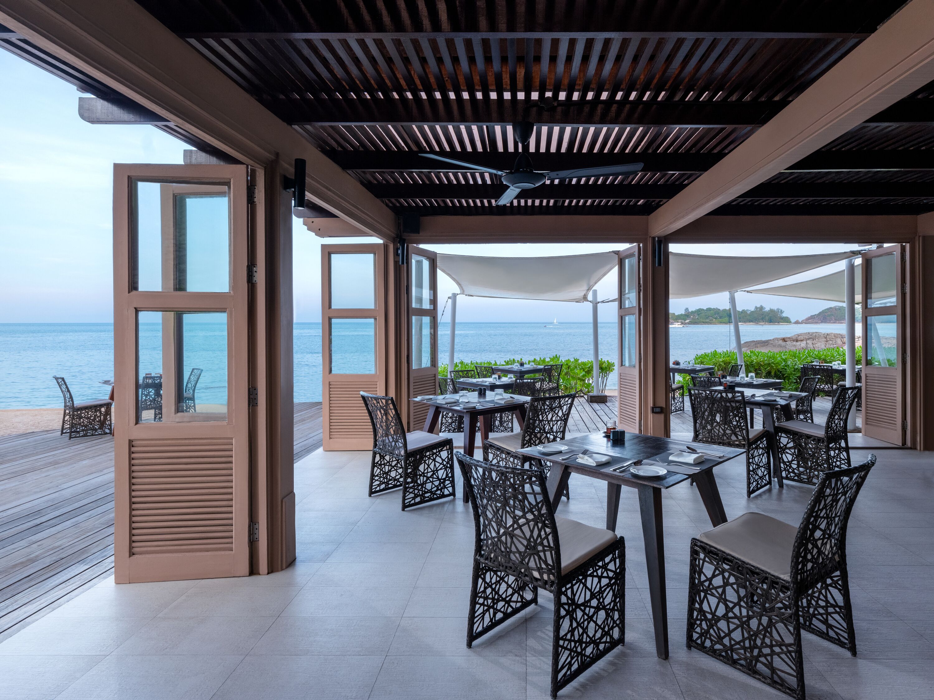 An outdoor dining area with tables and chairs, open doors leading to the sea, and a ceiling fan.