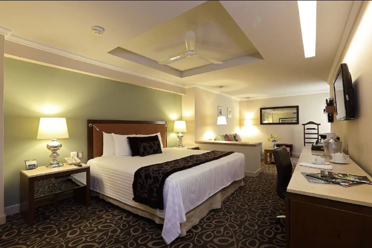 Habitación de hotel con una cama grande, dos mesitas de noche con lámparas, un escritorio con una silla, un televisor de pantalla plana montado en la pared y un ventilador de techo.