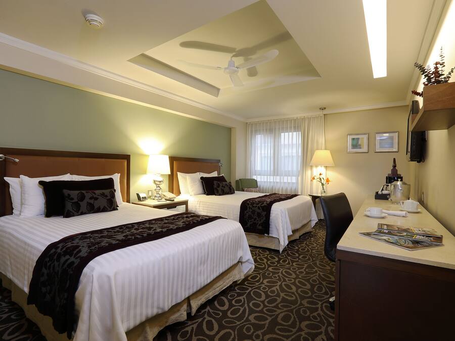 A hotel room with two beds, a desk, a chair, a ceiling fan, and a window with curtains.