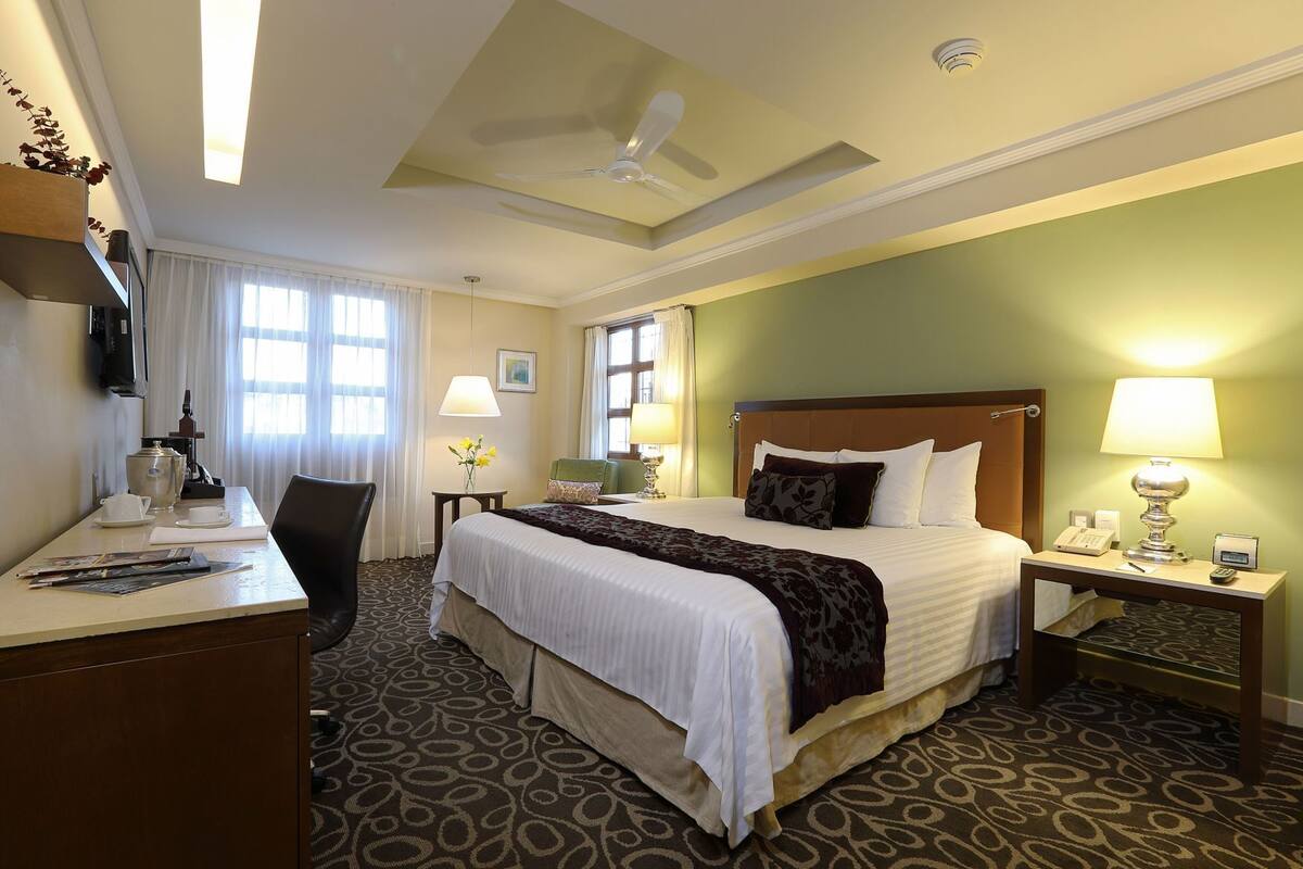 Habitación de hotel con cama, escritorio, silla, mesitas de noche, lámparas y ventilador de techo.