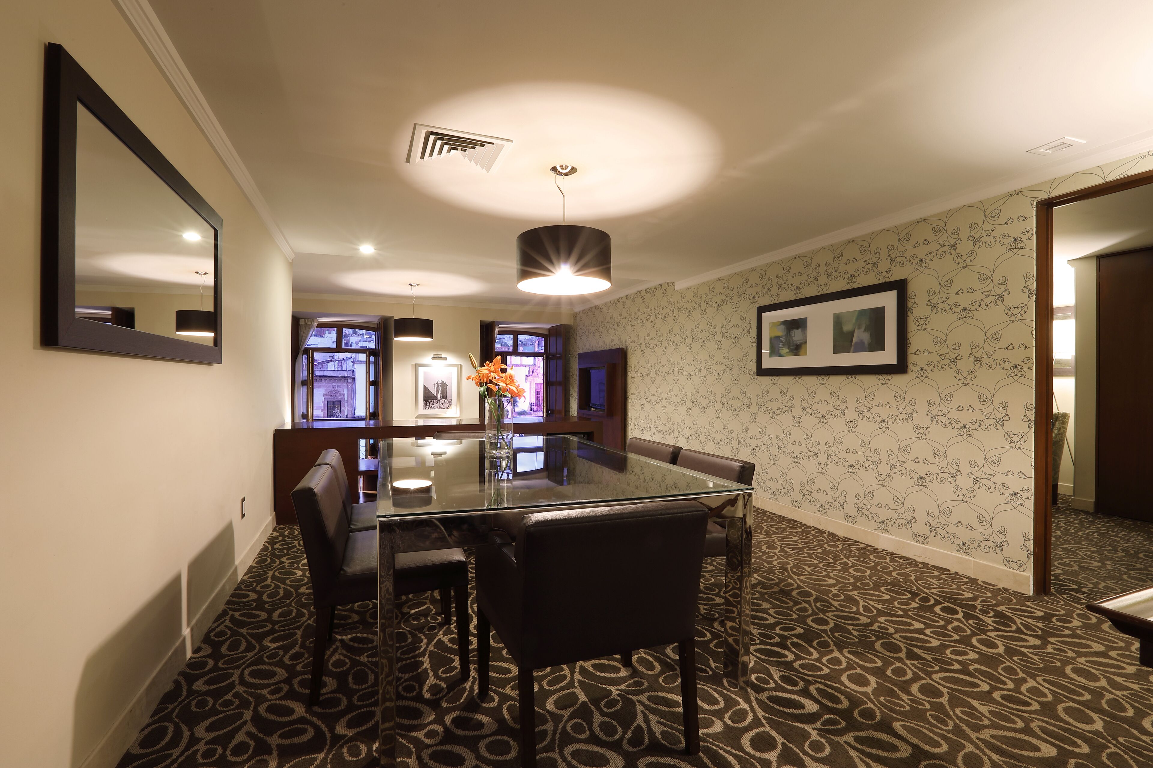 A dining area with a glass table, leather chairs, patterned carpet, and a wall-mounted mirror.